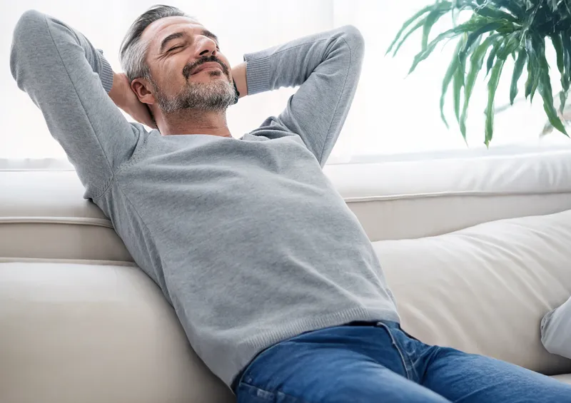 Entspannter Mann lehnt sich lächelnd auf einem Sofa zurück als Symbol für innere Ruhe, Wohlbefinden und ein ausgeglichenes Körpergefühl